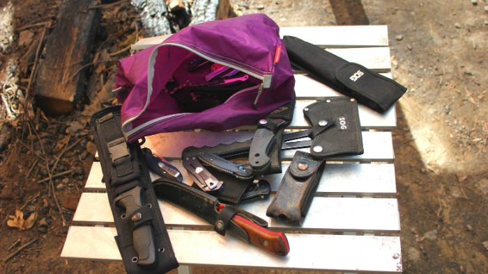 Different kinds of camping and survival knives on a table next to a purple bag.