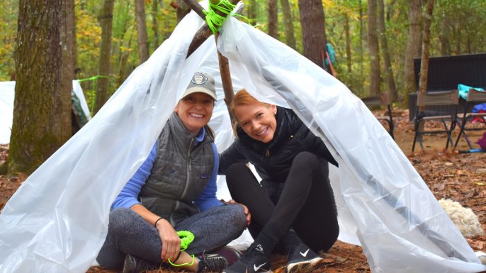 Two women sitting beneath a plastic tarp propped up with sticks during a survival training course in VA.