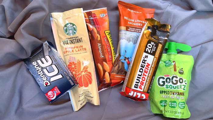Trail mix, granola bars, applesauce and other hiking snacks lying on a gray sleeping bag.