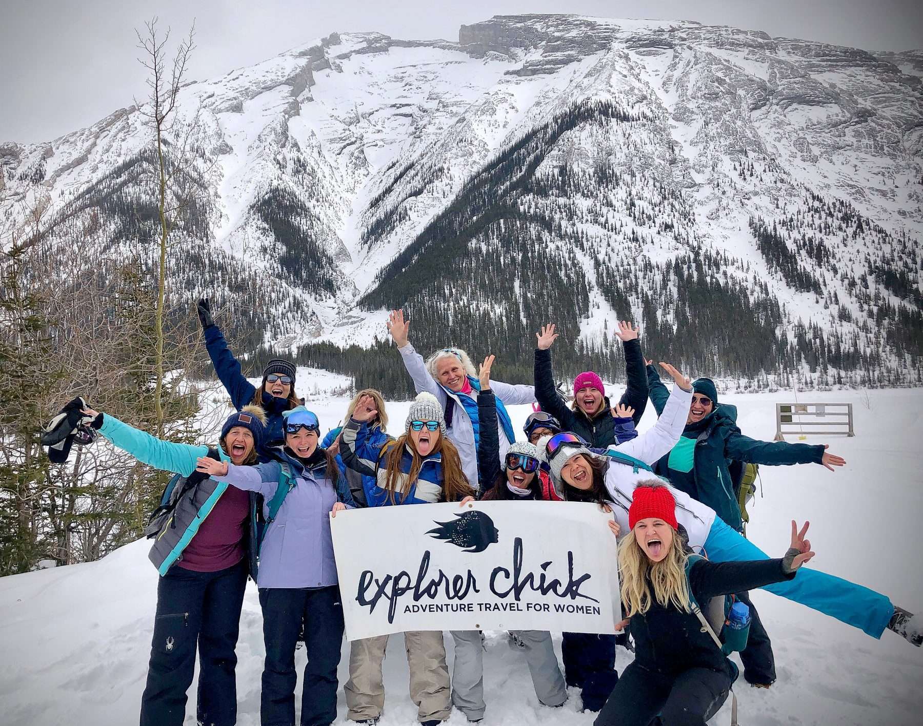 Women smiling and posing in front of a snow-covered mountain while holding an Explorer Chick banner