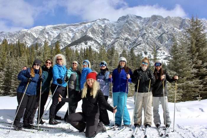 A small group of women on a guided tour, wearing skis and standing in front of a mountain and pine trees.