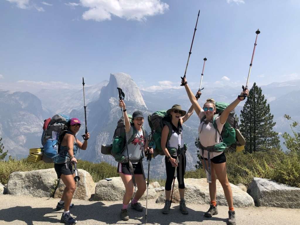 Women posing with backpacks and holding up trekking poles during a hike to Half Dome