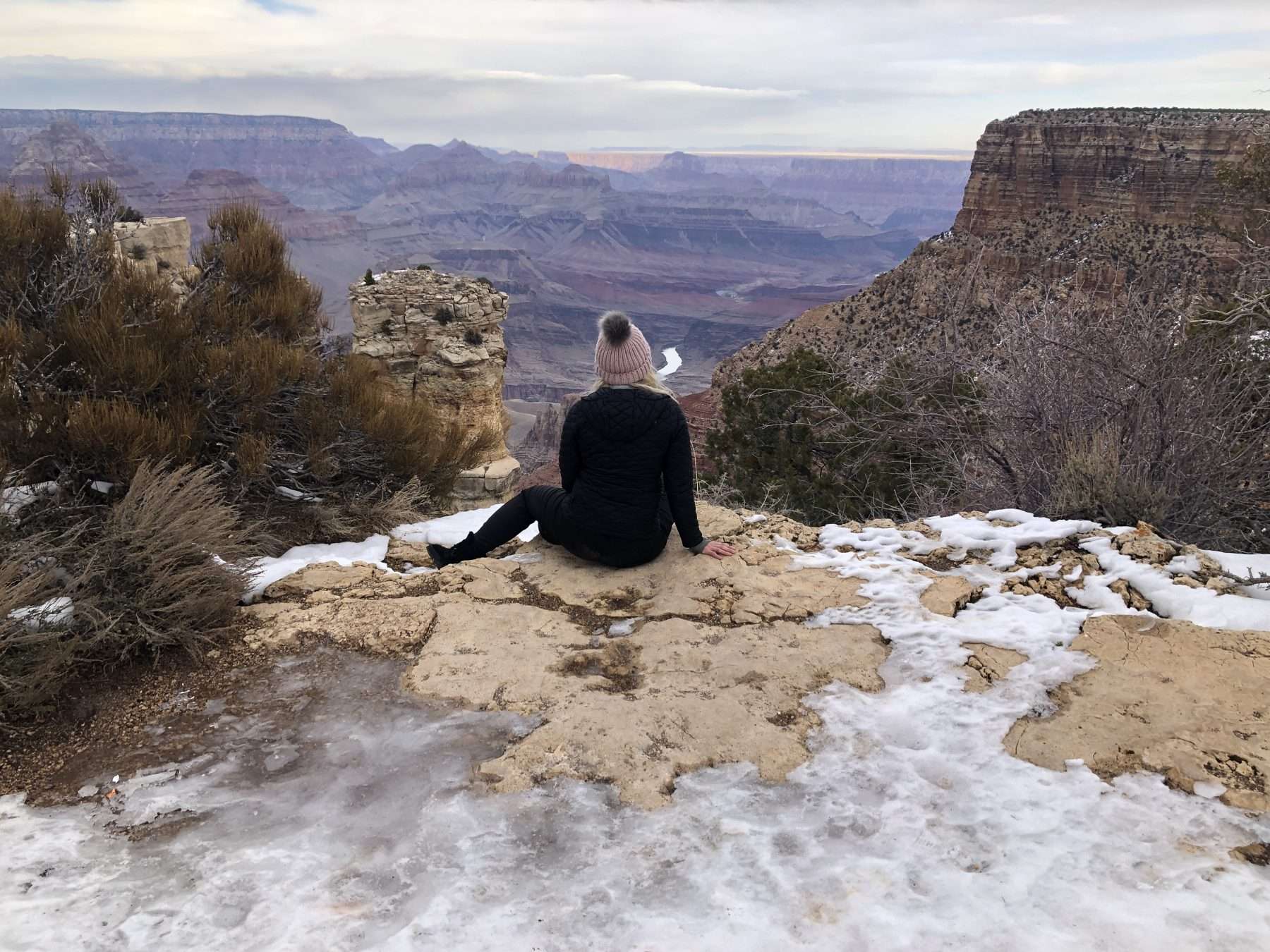Woman in black outfit and pink hat sitting on a rock surrounded by snow looking at the Grand Canyon