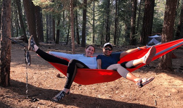 Two women in the woods smiling and sitting in a red camping hammock, a great gift idea for an outdoors-woman