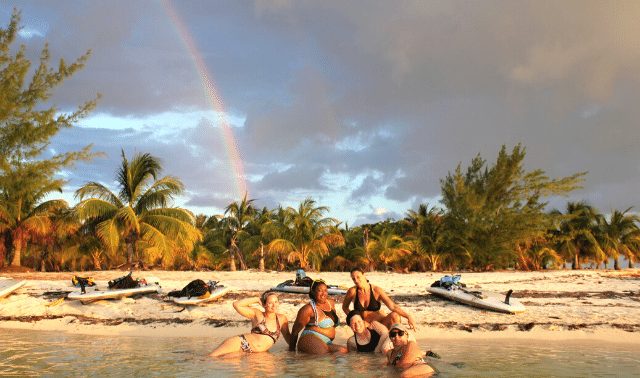 Five women traveling together posing on beach with palm trees and jet skis with a rainbow in the sky
