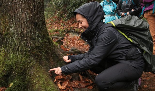 Woman in black coat leaning down and pointing to tree during a winter hike